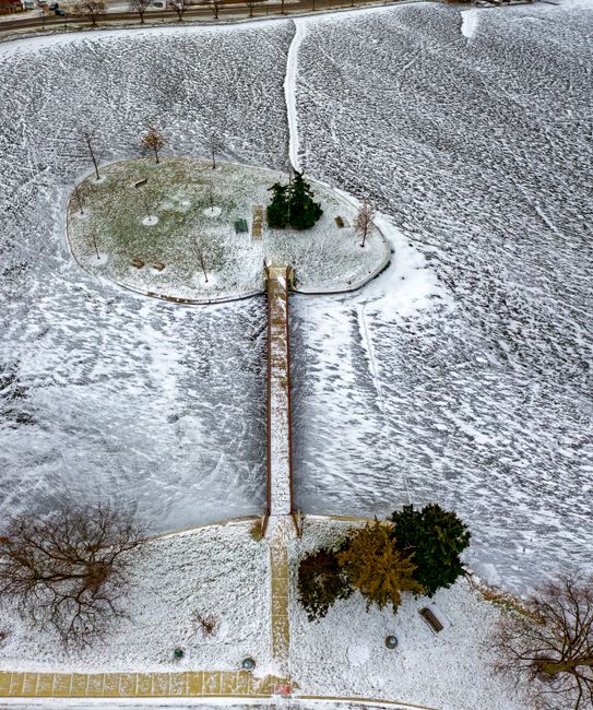 An aerial view of a small garden covered in a light layer of snow, showcasing a symmetrical layout divided by a central pathway leading to a circular, raised garden bed with a few small, leafless tree