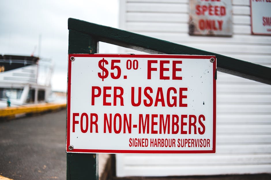A white rectangular sign with bold red text displaying a $5.00 fee per usage for non-members, attached to a dark green metal post with rounded edges. The background shows a grey pavement and part of a